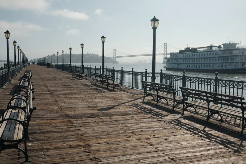 Empty wooden pier lined with benches and lamp posts, the Bay Bridge and a paddle-wheel riverboat in the background