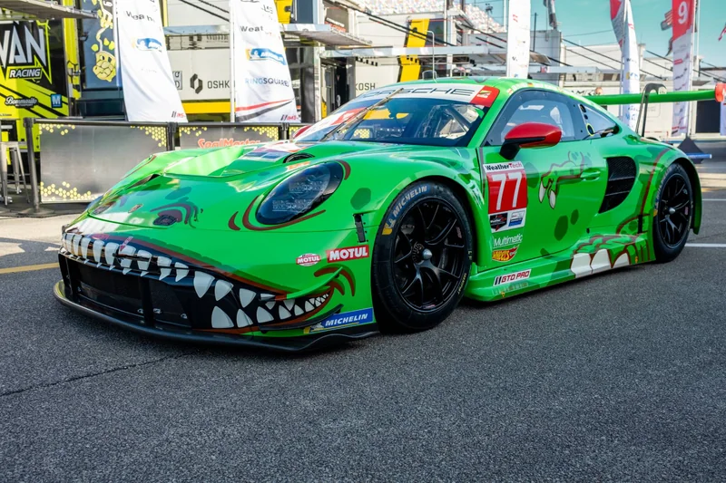 Bright green #77 Porsche 911 GT3 R race car with a cartoon dinosaur-teeth livery, parked in the paddock at Daytona International Speedway