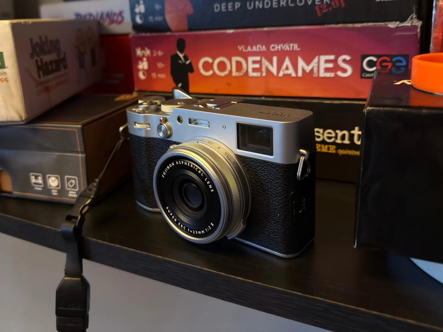 Silver and black Fujifilm X100V with its 23mm f/2 lens, sitting on a shelf in front of board game boxes